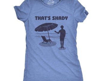 Puede incluir: Una camiseta de manga corta en azul jaspeado con un gráfico azul oscuro de una escena de playa, una tumbona bajo una sombrilla y una persona de pie. El texto "THAT'S SHADY" está impreso en letras azul oscuro sobre la ilustración.