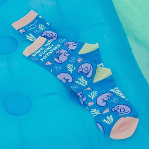 May include: A pair of blue socks with a pink trim and a pattern of manatees, seashells, and seaweed. The socks have the text "Save the Floaty Potatoes" printed on them.