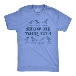 May include: A heather blue short-sleeve t-shirt featuring a dark blue graphic of seven bird species with the text "SHOW ME YOUR TITS" printed in the center. The birds are labeled as Blue Tit, Great Tit, Crested Tit, Coal Tit, Marsh Tit, Bearded Tit, and Long Tailed Tit.