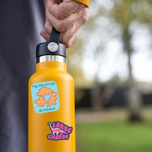 May include: A yellow water bottle with a black lid and handle is held by a person. The bottle has two stickers: one with orange dinosaur illustrations and the text "my favorite dinosaur", and another with the text "Large and in Charge".