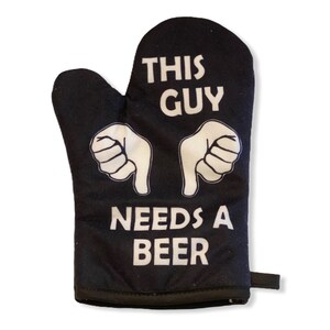May include: A black oven mitt featuring white text that reads THIS GUY NEEDS A BEER. The design includes two white graphic illustrations of hands with thumbs pointing downward toward the text. A small black fabric loop is attached to the side.