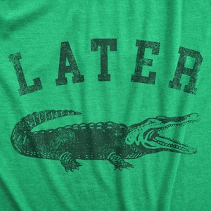 May include: Green t-shirt with the word "LATER" above a detailed illustration of an alligator. The alligator is dark green and has its mouth open. The text and graphic have a distressed, vintage look.