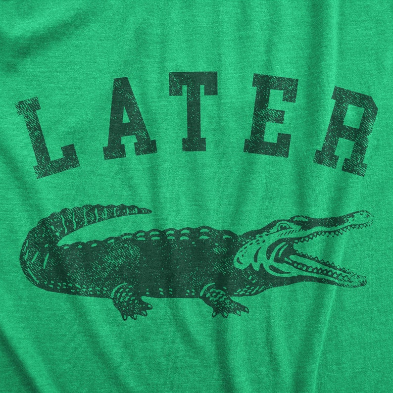 May include: Green t-shirt with the word "LATER" above a detailed illustration of an alligator. The alligator is dark green and has its mouth open. The text and graphic have a distressed, vintage look.
