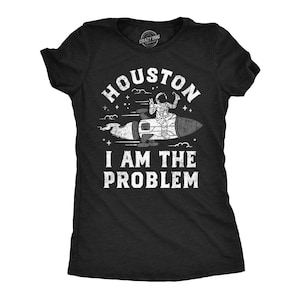 Può includere: Una t-shirt nera a maniche corte con una grafica bianca di un astronauta a cavallo di un razzo. Il testo recita HOUSTON I AM THE PROBLEM. Il design include stelle e nuvole, con una texture usurata sulla stampa grafica di colore bianco.