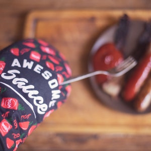 May include: A black oven mitt with red chili peppers and the text "Awesome Sauce" printed on it. The mitt is being used to hold a fork over a plate of food.