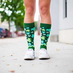 Może przedstawiać: Para białych trampek z zielonymi skarpetkami w wzór koniczyny. Skarpety sięgają do kolan i mają zielono-biało-niebieski kolor.