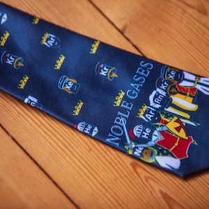May include: A dark blue necktie featuring a repeating pattern of small crowns and noble gas symbols including He, Ar, Kr, and Xe. The lower section displays the text NOBLE GASES above a group of illustrated figures wearing crowns and armor.