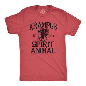 May include: A heathered red short-sleeve t-shirt featuring a black graphic of the character Krampus in the centre. The text printed in black ink reads KRAMPUS IS MY SPIRIT ANIMAL, with the word KRAMPUS arched above the figure and the rest below.