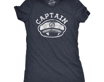 Peut inclure: Un t-shirt bleu marine foncé à manches courtes avec un motif blanc vieilli représentant une casquette de capitaine avec une ancre. Le mot CAPTAIN est imprimé en lettres blanches, grasses et arquées au-dessus de l'illustration.