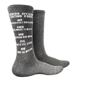 May include: A pair of charcoal gray crew socks featuring white printed text that reads: SENIOR CITIZEN TEXTING CODE: BFF BLADDER FAILS FREQUENTLY, OMG OLD, MOODY & GASSY, BRB BATHROOM RUN BREAK, TTYL TIME TO YELL LOUDLY, BTW BRING THE WALKER.