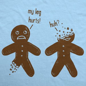 May include: Light blue t-shirt featuring a humorous illustration of two gingerbread men. One has a broken leg and says "my leg hurts!" The other has a broken head and says "huh?" The design is in brown.