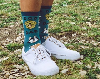 May include: Teal crew socks featuring a repeating pattern of dog faces and the text DOG DAD in dark blue lettering, paired with white lace-up sneakers. The socks display illustrations of golden retriever and husky faces with tongues out.