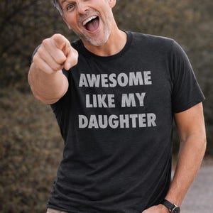 May include: A dark gray t-shirt with the text "AWESOME LIKE MY DAUGHTER" in white. The person is pointing at the viewer and wearing khaki pants and a black watch.