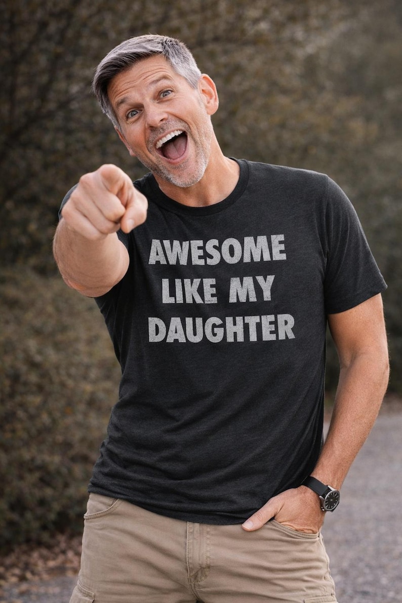 May include: A dark gray t-shirt with the text "AWESOME LIKE MY DAUGHTER" in white. The person is pointing at the viewer and wearing khaki pants and a black watch.