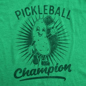 Puede incluir: Camiseta verde con un personaje de pepinillo sosteniendo una pala de pickleball y una pelota de pickleball. El texto "Pickleball Champion" est&aacute; impreso en la camiseta.