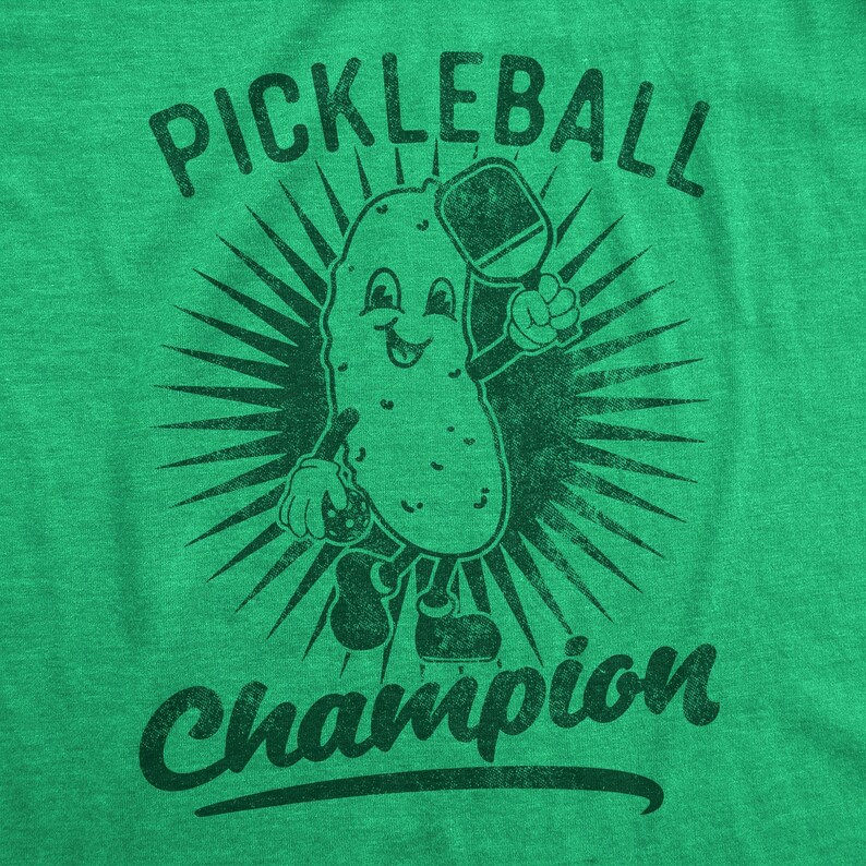 Puede incluir: Camiseta verde con un personaje de pepinillo sosteniendo una pala de pickleball y una pelota de pickleball. El texto "Pickleball Champion" est&aacute; impreso en la camiseta.
