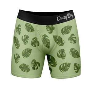 Puede incluir: Calzoncillos tipo bóxer de color verde claro con un patrón repetitivo de hojas de monstera de color verde oscuro. La prenda tiene una cintura elástica ancha de color negro con el texto Crazy Dog en blanco.