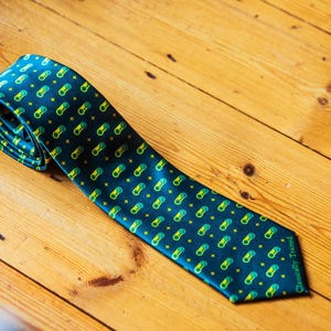May include: A dark green necktie featuring a repeating pattern of small yellow and light green video game controllers and stars. The bottom corner of the tie includes the text Classically Trained printed in a small, light green font.