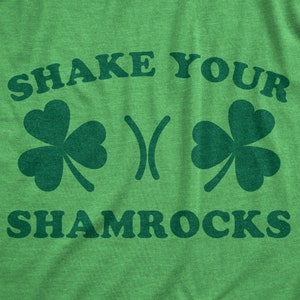 以下が含まれることがあります： 黒い文字で「Shake Your Shamrocks」と書かれた緑色のTシャツ。Tシャツには、クローバーのデザインが2つあります。