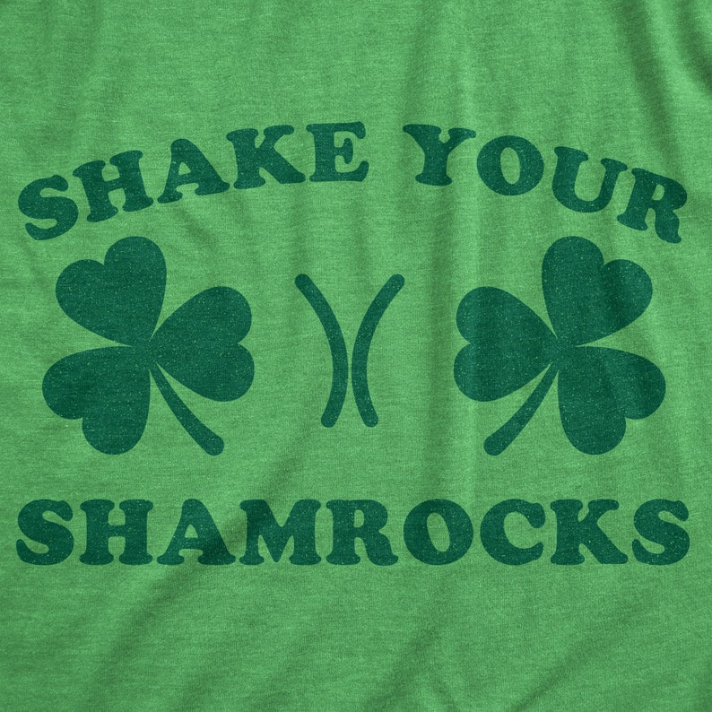 以下が含まれることがあります： 黒い文字で「Shake Your Shamrocks」と書かれた緑色のTシャツ。Tシャツには、クローバーのデザインが2つあります。
