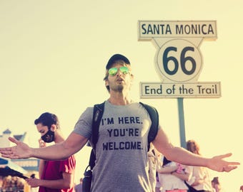 May include: A grey short-sleeved t-shirt with the black text I'M HERE. YOU'RE WELCOME. worn by a person with a black baseball cap and reflective sunglasses, standing in front of a Santa Monica Route 66 End of the Trail sign.