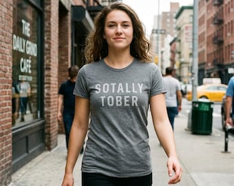 Puede incluir: Una camiseta gris jaspeado de manga corta con el texto blanco SOTALLY TOBER impreso en una fuente sans-serif clara en el pecho. La prenda tiene cuello redondo y un corte estándar, confeccionada con un tejido suave y texturizado.