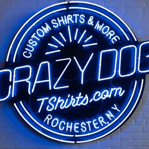 May include: A blue neon sign for Crazy Dog T-shirts. The sign is circular with the words "Custom Shirts & More" and "Rochester, NY" in a circle around the words "Crazy Dog" and "TShirts.com".