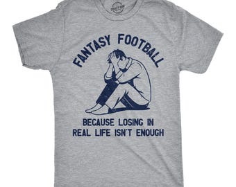 Op de afbeelding: Een grijs gemêleerd t-shirt met korte mouwen en een donkerblauwe afbeelding van een persoon die zittend het hoofd in de handen houdt. De tekst luidt FANTASY FOOTBALL BECAUSE LOSING IN REAL LIFE ISN'T ENOUGH in een verweerd marineblauw lettertype.