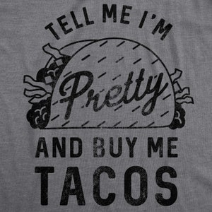 May include: A gray t-shirt with black text that reads "Tell me I'm pretty and buy me tacos". The text is stylized to look like a vintage design. A black and white illustration of a taco is in the center of the text.