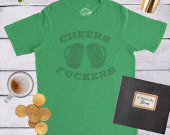 May include: A green short-sleeve t-shirt featuring a dark green graphic of two beer mugs and the text "CHEERS FUCKERS" centered on the chest. The shirt is displayed on a light wood surface alongside gold coins, a glass of beer, and a black book.