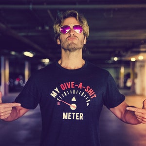 以下が含まれることがあります： ピンクのサングラスをかけ、黒いTシャツを着た男性。Tシャツには「My Give-A-Shit Meter」という文字と燃料計のグラフィックが描かれている。
