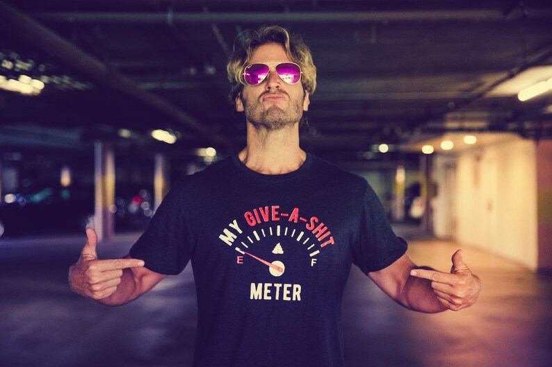以下が含まれることがあります： ピンクのサングラスをかけ、黒いTシャツを着た男性。Tシャツには「My Give-A-Shit Meter」という文字と燃料計のグラフィックが描かれている。