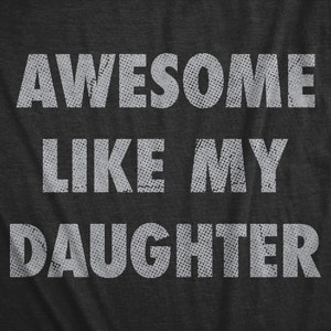 May include: Dark gray t-shirt with the text "AWESOME LIKE MY DAUGHTER" in white, distressed lettering. The text is centered and takes up most of the shirt's surface. The font has a dotted texture, giving it a vintage look.