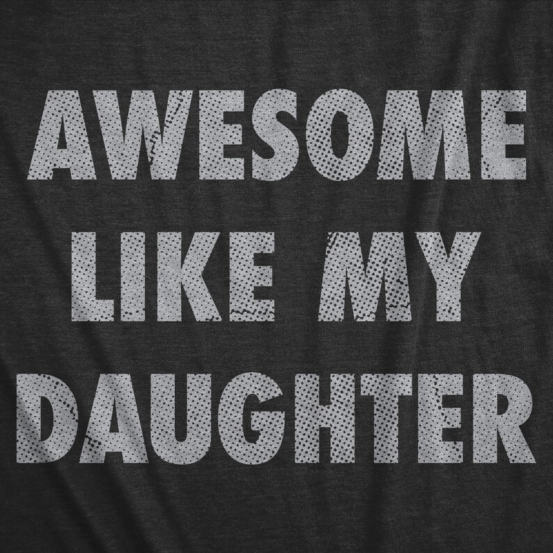 May include: Dark gray t-shirt with the text "AWESOME LIKE MY DAUGHTER" in white, distressed lettering. The text is centered and takes up most of the shirt's surface. The font has a dotted texture, giving it a vintage look.