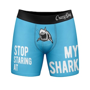 Puede incluir: Calzoncillos tipo bóxer de color azul claro con una cinturilla negra que incluye el texto Crazy Dog. La parte delantera muestra un gráfico de un tiburón con la boca abierta y el texto blanco STOP STARING AT MY SHARK.