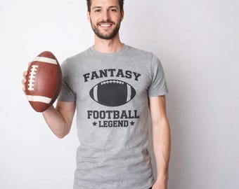 Op de afbeelding: Een grijs gemêleerd T-shirt met korte mouwen en een zwarte grafische opdruk met de tekst FANTASY FOOTBALL LEGEND en een illustratie van een football in het midden. Het shirt wordt gecombineerd met een bruine leren football met witte veters.