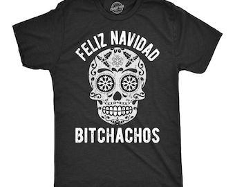 May include: A charcoal gray short-sleeve t-shirt featuring a white graphic of a sugar skull with snowflake eye sockets. The text FELIZ NAVIDAD is printed in an arch above the skull, and the word BITCHACHOS is printed in white below the skull.