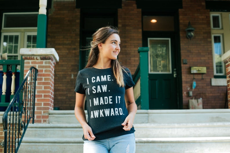 K&ouml;nnte beinhalten: Eine Frau tr&auml;gt ein dunkelgraues T-Shirt mit dem Text "I came. I saw. I made it awkward." in wei&szlig;en Buchstaben. Sie steht auf einer Treppe vor einem Haus.