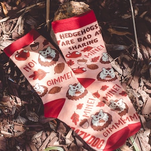 May include: A pair of pink socks with a red cuff. The socks feature a hedgehog design with the text "Hedgehogs are bad at sharing mine!" and "Gimme! Mine! Gimme! Mine!"