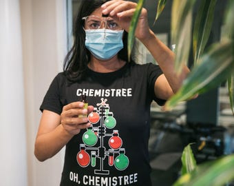 Pode incluir: Uma t-shirt preta com um gráfico de uma árvore de Natal construída com material de laboratório, incluindo balões de fundo redondo vermelhos e verdes e tubos de ensaio. O texto diz CHEMISTREE no topo e OH, CHEMISTREE na base.