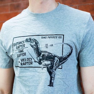 Könnte beinhalten: Ein graues, meliertes Kurzarm-T-Shirt mit einem schwarzen Grafikdruck eines Velociraptors. Das Design enthält den Text DINO PHYSICS 101, eine physikalische Gleichung sowie die Wörter DISTANCE RAPTOR geteilt durch TIME RAPTOR gleich VELOCI RAPTOR.
