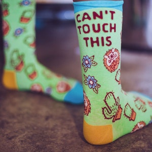 May include: A pair of green socks with a colorful pattern of flowers, cacti, and hedgehogs. The socks have the text "Can't Touch This" written on them.