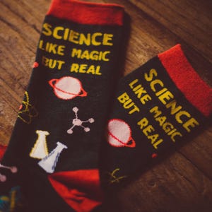 May include: A pair of black socks with red cuffs and heels, featuring the yellow text SCIENCE LIKE MAGIC BUT REAL. The socks are decorated with colorful scientific graphics including a planet with rings, a molecule, and laboratory beakers.