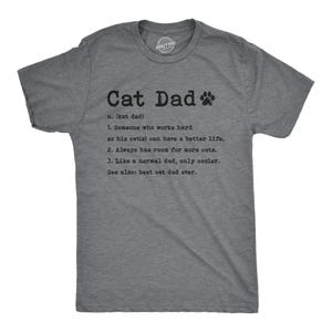 May include: A heather gray short-sleeve t-shirt featuring black text that reads: Cat Dad n. (kat dad) 1. Someone who works hard so his cat(s) can have a better life. 2. Always has room for more cats. 3. Like a normal dad, only cooler. See also: best cat dad ever.