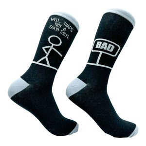 May include: A pair of black socks with white text and illustrations. One sock says "Well that's not a good sign." and has a stick figure illustration. The other sock says "BAD" with a road sign illustration.
