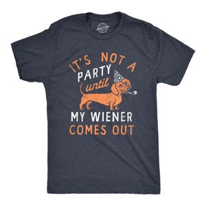 May include: A dark charcoal grey short-sleeve t-shirt featuring a graphic of an orange dachshund wearing a party hat and holding a party blower. The text reads: IT'S NOT A PARTY UNTIL MY WIENER COMES OUT. The design is printed in orange and white.
