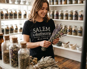 Puede incluir: Una camiseta negra de manga corta con texto blanco que dice SALEM Apothecary POTIONS TONICS ELIXIRS est. 1690. La prenda se lleva puesta mientras se sostiene un manojo de lavanda seca y otras hierbas en una tienda llena de frascos de vidrio.