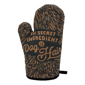 Pode incluir: Uma luva de forno preta com um padrão de pelo castanho e o texto THE SECRET INGREDIENT IS Dog Hair impresso no centro. A luva tem uma secção para o polegar à esquerda e uma pequena alça de tecido preto no canto inferior direito.