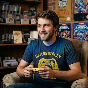 Könnte beinhalten: Ein kurzärmeliges T-Shirt in meliertem Marineblau mit einer gelben Grafik von Händen, die einen Videospiel-Controller halten, und dem darüber gedruckten Text "CLASSICALLY". Das T-Shirt besteht aus einem weichen, leichten Stoff.