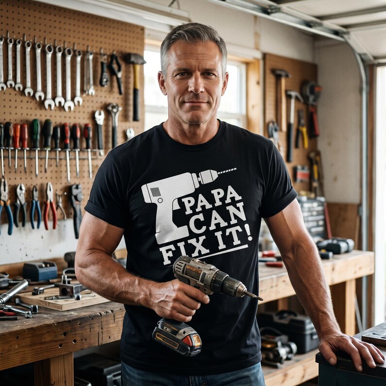 May include: A black t-shirt with the text "PAPA CAN FIX IT!" and a graphic of a drill. The person in the image is holding a cordless drill. Tools are visible on the pegboard in the background.
